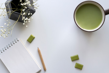 Matcha chocolate, brown cup with green milk tea, notebook, pencil, polygonal grey vase with little white flowers on the light background. Top view, flat lay, copy space, mock up