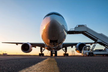 Naklejka premium Widok z przodu samolotu pasażerskiego białego szerokokadłubowego ze schodami na pokład na płycie lotniska w wieczornym słońcu