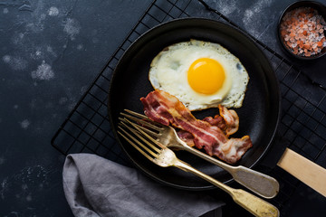Traditional English breakfast with fried eggs and bacon in cast iron pan on dark concrete background. Top view.