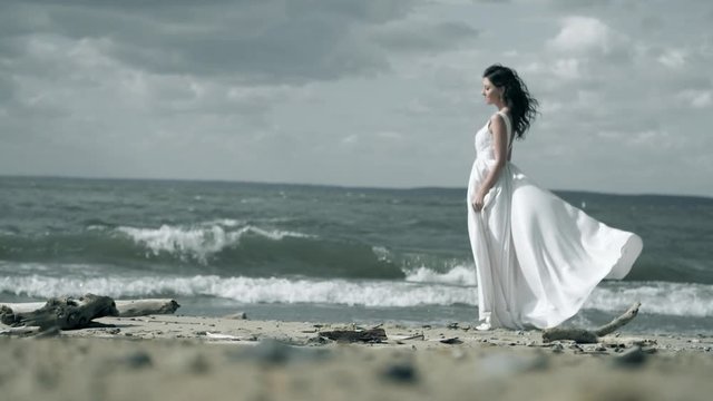 Lonely beautiful woman in a white dress stands on the ocean. Girl is holding a dress that flutters in wind slow motion. 
