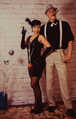 a girl with short black hair with a bandage with sequins and feathers on her head in a dress with sequins in the Chicago style of 20 years, with a man in a white shirt with suspenders and a black hat