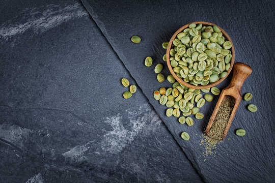 Raw green unroasted coffee in wooden bowl with ground beans in wood scoop on black slate board