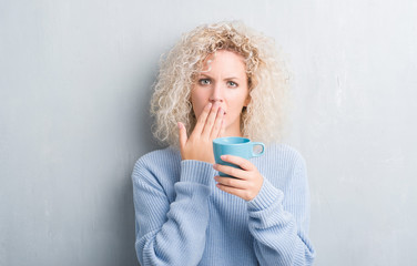 Young blonde woman over grunge grey wall driking a cup of coffee cover mouth with hand shocked with shame for mistake, expression of fear, scared in silence, secret concept