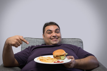 Obese man lying on sofa happily pointing at plate with burger and french fries in his hand