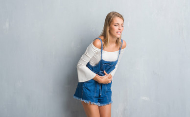 Beautiful young woman standing over grunge grey wall with hand on stomach because nausea, painful...