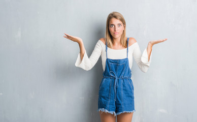 Beautiful young woman standing over grunge grey wall clueless and confused expression with arms and...