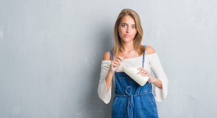 Beautiful young woman over grunge grey wall holding bottle of fresh milk with a confident expression on smart face thinking serious