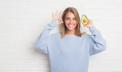 Beautiful young woman over white brick wall eating fresh avocado doing ok sign with fingers, excellent symbol