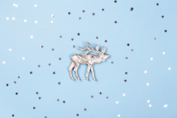 Obraz na płótnie Canvas Christmas figurine of reindeer and silver confetti on blue background. Minimal New Year card, top view.
