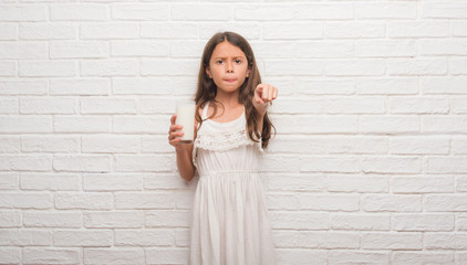 Young hispanic kid over white brick wall drinking a glass of milk pointing with finger to the camera and to you, hand sign, positive and confident gesture from the front