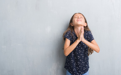 Young hispanic kid over grunge grey wall begging and praying with hands together with hope expression on face very emotional and worried. Asking for forgiveness. Religion concept.