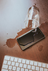 Smartphone and keyboard wet by accident on wooden table. Glass of water spilled.