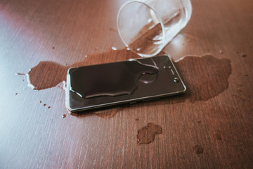 Smartphone wet by accident on wooden table. Glass of water spilled.