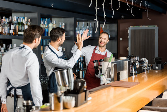 handsome barmen in aprons high five at workplace