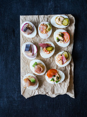Stuffed eggs with various topping. Prosciutto, herring, pickled cucumber, sausage, radish, tomato, capers and sesame on boiled egg halves. Top view