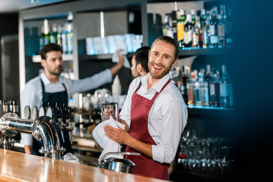 Smiling Barman Polishing Glass With Cloth At Wooden Counter At Bar