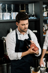 handsome barman in apron preparing cocktail in glass with shaker