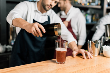 adult barman in apron pouring cocktail in glass from shaker
