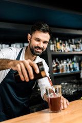 smiling barman pouring cocktail from shaker in glass at wooden counter