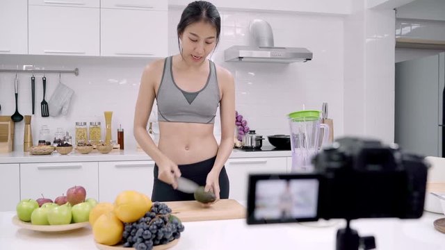 Blogger sporty Asian woman using camera recording how to make avocado juice video for her subscriber, female use organic fruit making avocado juice by herself at home. Healthy food concept.
