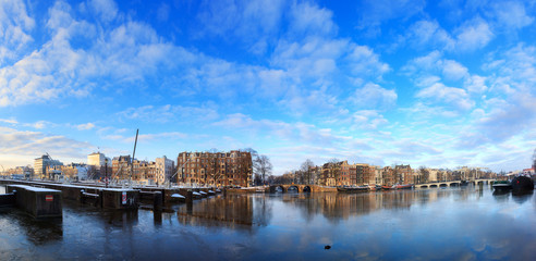Fototapeta premium Beautiful 180 degree cityscape panorama of the river Amstel in Amsterdam, the Netherlands, in winter with ice and snow with the famous skinny bridge (magere brug)