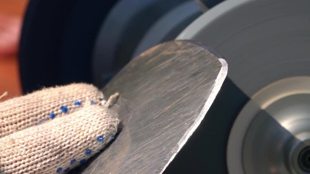 Sharpening of worn garden tool with unbalanced grinding machine in slow motion close up. Workshop background with fingers in safety glove, round-pointed steel shovel and turning grindstone wheel.