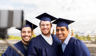 education, graduation, technology and people concept - group of happy international male students in mortar boards and bachelor gowns taking picture by smartphone selfie stick outdoors