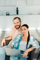 handsome husband and wife sitting on couch, looking at camera and using laptop at home