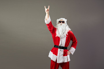 Fototapeta na wymiar Santa Claus with a long white beard in sunglasses and headphones shows a rock sing on the gray background.