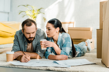 handsome husband and wife lying on floor, drinking coffee and working on blueprints of new house at home, moving concept
