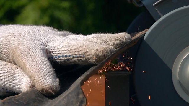 Sharpening of soiled garden tool with unbalanced grinding machine in slow motion close up. Panning of traditional machinery scene with detailed round-pointed steel shovel, waving hand in safety glove.