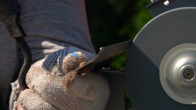 Sharpening of soiled garden tool with unbalanced grinding machine in slow motion close up. Panning of traditional machinery scene with detailed hoe, waving hand in safety glove and turning grindstone.