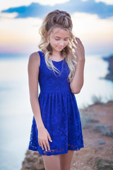 Cute young lady with curly blond hair posing on beach wearing tender blue dress on sea side background and blue heaven sky. Adore scene of pretty emancipated girl with tender skin.