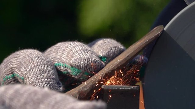 Sharpening of old grange ax with unbalanced grinding machine in slow motion close up. Traditional machinery scene with retro tool, waving hand in safety glove, turning grindstone wheel and sparks.