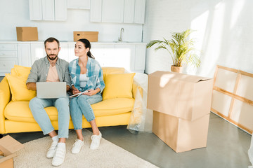 Fototapeta na wymiar concentrated couple sitting on couch, using laptop and planning relocation to new house, moving concept