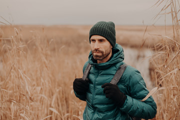People and tourism concept. Pensive unshaven handsome young man with rucksack, dressed in stylish clothes, focused into distance, poses against outdoor field background. Lifestyle. Adventure