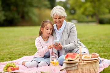 family, leisure and technology concept - happy grandmother and granddaughter with smartphone having picnic at summer park