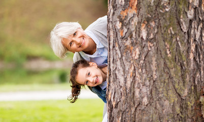 family, leisure and people concept - happy grandmother and granddaughter peeking out tree at summer park - obrazy, fototapety, plakaty