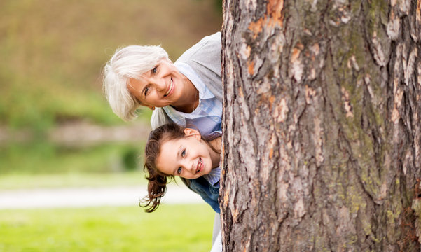 family, leisure and people concept - happy grandmother and granddaughter peeking out tree at summer park
