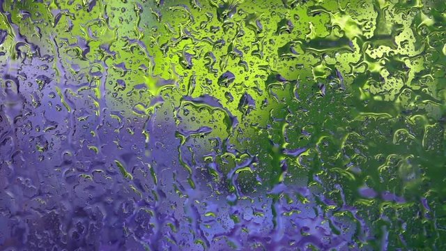 Green and blue defocused view through rainy window close up. Water shower on vertical flat transparent glass in slow motion. Amazing abstract natural background with vibrant aqua texture. High speed.