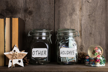 Two piggy banks, with different amounts of money for holidays, stand on a wooden bookshelf.