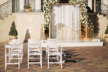 Weddind decoration on open air. Floral decor of a beautiful white and gold arch. Beautiful beckground view of restaurant and trees. Stool for ceremony