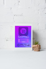 digital tablet with shopping application and green potted plant near white brick wall