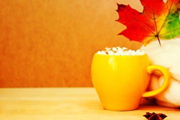 Cozy autumn concept with yellow cup of cocoa with marshmallow and cinnamon, autumn leaves and knitted white scarf on wooden background. Copy space