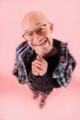 Funny portrait from above of unusual crazy bald man in glasses shows different emotions. Facial expression. Stylish boy looking up in fashionable tracksuit with flowers pattern on pink background.