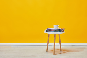 Little wooden table with pile of magazines and white cup of coffee near yellow wall - Powered by Adobe