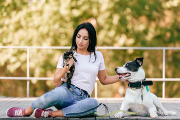 Lifestyle portrait of young adorable brunette girl with little cat and big hound dog sitting outdoor in park. Happy cheerful smiling teen hugging pets. Positive female with  hunting puppy and pussycat