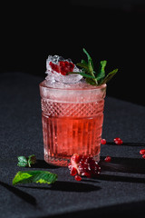 Cold cocktail with ice and garnet seeds, piece of pomegranate behind on dark background