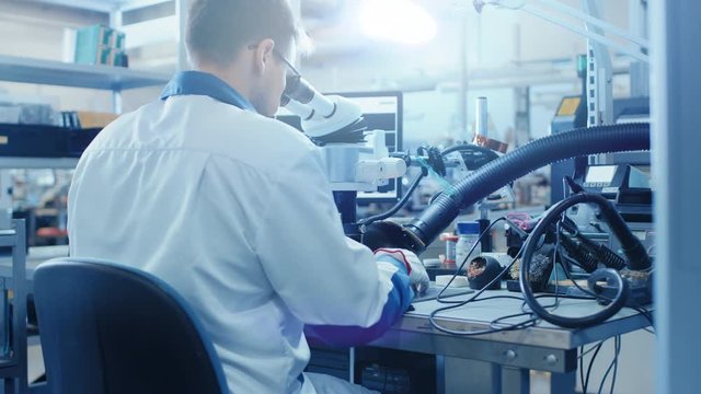 Electronics Factory Worker in White Work Coat Inspects a Printed Circuit Board Through a Digital Microscope. High Tech Factory Facility.