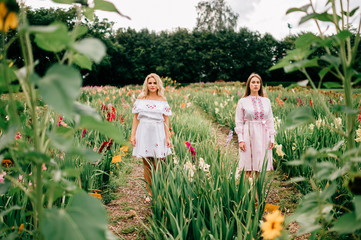 Two pretty young female friends in ethnic dresses relaxing in flower garden in summer. Couple of...